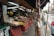 A bustling street market with various colorful fruits displayed on a series of red counters. The market is sheltered by a tarp, and the street outside is lined with signs and passing vehicles. A variety of fruits including bananas, pineapples, and dragon fruits are visible.