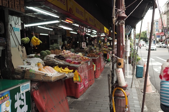 A bustling street market with various colorful fruits displayed on a series of red counters. The market is sheltered by a tarp, and the street outside is lined with signs and passing vehicles. A variety of fruits including bananas, pineapples, and dragon fruits are visible.