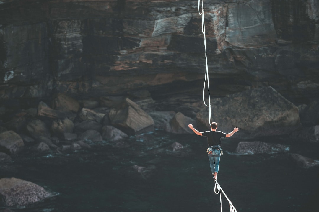 The photo shows the back of a person who is wearing a black t-shirt and jeans and is walking a tight rope over a river towards a steep rock face.