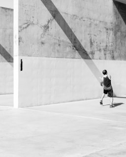 A man in athletic clothing is playing handball on a concrete court. Strong diagonal shadows cast across the textured wall, creating a geometric pattern. The scene is in black and white, emphasizing the contrast and shadows.