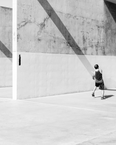 A man in athletic clothing is playing handball on a concrete court. Strong diagonal shadows cast across the textured wall, creating a geometric pattern. The scene is in black and white, emphasizing the contrast and shadows.