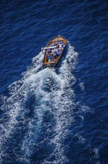 A small wooden boat travels through deep blue ocean waters, leaving trails of white foam in its wake. Two people are visible on the boat, engaged in leisurely activities.