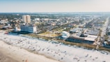 A beachside cityscape featuring a wide expanse of sandy beach dotted with people and umbrellas, adjacent to a town with a mix of residential and commercial buildings. A large hotel stands prominently near the shoreline, and the city extends into the distance with dense greenery and urban infrastructure.