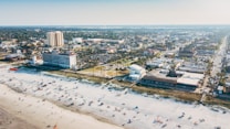 A beachside cityscape featuring a wide expanse of sandy beach dotted with people and umbrellas, adjacent to a town with a mix of residential and commercial buildings. A large hotel stands prominently near the shoreline, and the city extends into the distance with dense greenery and urban infrastructure.