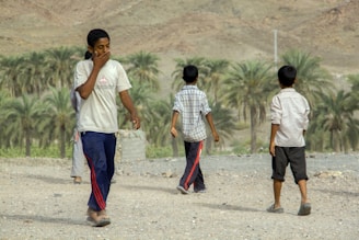 Several young boys are walking on a rocky path in what appears to be a rural or semi-rural area. Three of them have their backs to the camera, and one of them is covering his mouth with his hand. In the background, there are palm trees and arid mountains.