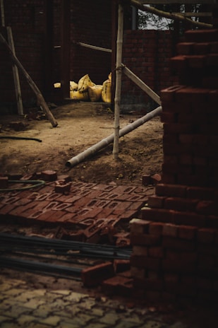 A construction site with stacks of red bricks and several yellow bags on a dirt ground, surrounded by wooden poles. The bricks are neatly piled, and the bags appear to be filled with construction materials.