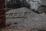 A pile of gravel covers a section of ground with a wooden ladder laid across it. Surrounding the pile, there are stray bricks scattered on a paved surface. A dilapidated wall with visible wear and chipping forms the backdrop alongside some overgrown vegetation.