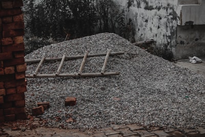 A pile of gravel covers a section of ground with a wooden ladder laid across it. Surrounding the pile, there are stray bricks scattered on a paved surface. A dilapidated wall with visible wear and chipping forms the backdrop alongside some overgrown vegetation.