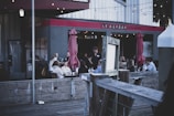 A group of people are dining in an outdoor area of a restaurant named 'Le Garage'. The setting appears cozy with string lights hanging above and red umbrellas providing shade. The atmosphere suggests evening dining, with people engaged in conversation and a server attending to a table.