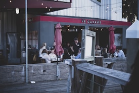 A group of people are dining in an outdoor area of a restaurant named 'Le Garage'. The setting appears cozy with string lights hanging above and red umbrellas providing shade. The atmosphere suggests evening dining, with people engaged in conversation and a server attending to a table.
