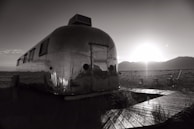 A vintage metal trailer with a reflective surface stands in an open field, with the sun setting behind distant mountains. A wooden path leads to the trailer, and an empty metal chair is positioned nearby. The scene captures a serene and nostalgic atmosphere.