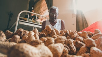 An elderly man is sitting beside a large pile of peanuts. He is wearing glasses and a white tank top. In the background, another person can be seen on a bed, with light coming through a window covered by curtains.