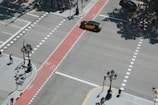 An aerial view of a city intersection with clearly marked pedestrian crossings and bike lanes. Several people are waiting at corners, while a yellow and black taxi is moving through the intersection. Surrounding the intersection are lamp posts and traffic signs. Shadows of nearby trees cast on the pavement indicate a sunny day.
