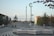 A public urban space featuring a unique design with white curved lines on a dark surface, creating an optical illusion of movement. People are walking and standing in the area, which includes a small hill or mound. There is a decorative lamp post with a crescent moon symbol and a square water fountain with streams of water.