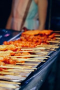 Grilled skewers of meat arranged in rows on a tray, each stick uniformly placed. A person in the background appears out of focus, suggesting a busy market or street food setting with dim lighting.