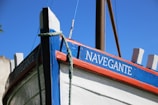 A close-up view of a boat's bow with a vibrant painting. The boat's name, 'Navegante', is clearly visible in white letters on a blue planking. The hull is painted white with red and blue accents, and a thick rope is draped over the side. The mast of the boat is visible in the background against a clear blue sky.
