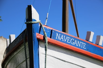 A close-up view of a boat's bow with a vibrant painting. The boat's name, 'Navegante', is clearly visible in white letters on a blue planking. The hull is painted white with red and blue accents, and a thick rope is draped over the side. The mast of the boat is visible in the background against a clear blue sky.