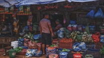 A bustling market stall is filled with various fresh vegetables and fruits, including tomatoes, radishes, green beans, cucumbers, and watermelons. Two people are present, one standing and the other sitting, possibly engaged in a transaction. The produce is organized in crates and bags, with a colorful display. The stall is covered with a canopy, and other market goods are visible in the background.