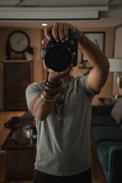 A person is holding a large camera, taking a self-portrait in a room. The room features soft lighting, with a clock on the wall and a couch visible in the background. The person is wearing casual clothing, and several bracelets can be seen on their arm.