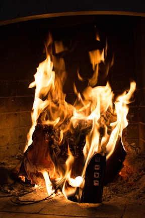 A lit fireplace contains a burning object, which appears to be a piece of electronic equipment with visible social media icons. The flames are vivid, casting a warm glow over the scene.