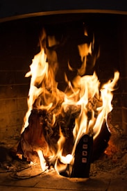 A lit fireplace contains a burning object, which appears to be a piece of electronic equipment with visible social media icons. The flames are vivid, casting a warm glow over the scene.