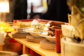 A vibrant street food market scene featuring a variety of skewered foods arranged neatly on ceramic plates. The skewers hold various types of grilled items, likely including sausages and other meats. The warm lighting gives the scene a cozy, inviting ambiance.