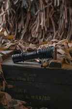 A black tactical flashlight is prominently placed on a wooden crate surrounded by spent bullet casings. The background consists of a draped camouflage net, suggesting a military or tactical context. The lighting is subdued, creating a somber atmosphere.