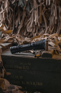 A black tactical flashlight is prominently placed on a wooden crate surrounded by spent bullet casings. The background consists of a draped camouflage net, suggesting a military or tactical context. The lighting is subdued, creating a somber atmosphere.