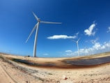 Tall wind turbines stand on a sandy landscape under a clear blue sky with scattered clouds. The terrain is open and expansive, with sparse vegetation and patches of water.