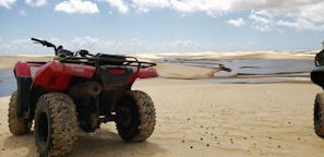 A red all-terrain vehicle (ATV) is parked on a sandy landscape near small water bodies, under a partly cloudy sky. The dunes extend into the distance, creating a vast and open desert-like panorama.
