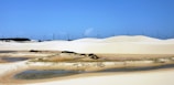 A vast sandy desert with gentle undulating dunes forms the foreground, dotted with patches of sparse vegetation and shallow pools of water. In the background, a row of wind turbines lines the horizon under a clear blue sky, suggesting a juxtaposition of natural and technological elements.