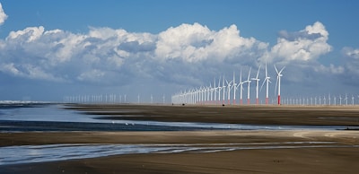 A vast landscape featuring a long line of wind turbines stretching into the distance. The scene captures a wide expanse of sandy beach with shallow water pools reflecting the blue sky and fluffy white clouds above. The horizon showcases numerous wind turbines, emphasizing renewable energy in a coastal environment.
