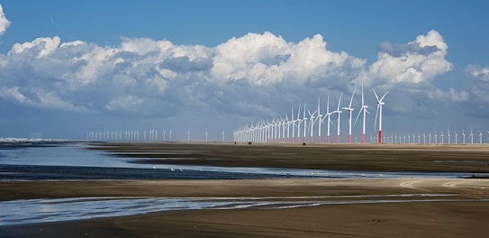 A vast landscape featuring a long line of wind turbines stretching into the distance. The scene captures a wide expanse of sandy beach with shallow water pools reflecting the blue sky and fluffy white clouds above. The horizon showcases numerous wind turbines, emphasizing renewable energy in a coastal environment.