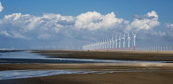 A vast landscape featuring a long line of wind turbines stretching into the distance. The scene captures a wide expanse of sandy beach with shallow water pools reflecting the blue sky and fluffy white clouds above. The horizon showcases numerous wind turbines, emphasizing renewable energy in a coastal environment.