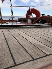 The image features a wooden deck of a boat with visible grain lines, surrounded by a metal railing. A red lifebuoy is mounted near the rear, and the sky is partly cloudy as the sea stretches out in the background. Various items, like ropes and equipment, are placed on the deck.
