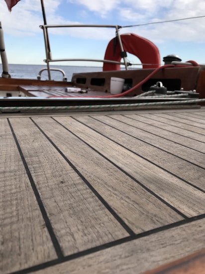 The image features a wooden deck of a boat with visible grain lines, surrounded by a metal railing. A red lifebuoy is mounted near the rear, and the sky is partly cloudy as the sea stretches out in the background. Various items, like ropes and equipment, are placed on the deck.