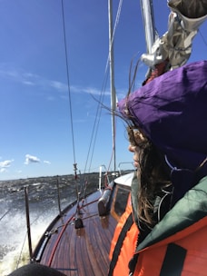 A person wearing a bright orange and green jacket with a purple hood is on a wooden sailboat. The boat is moving through choppy waters under a clear blue sky. There is a sense of motion as the boat cuts through the waves, and the person's hair blows in the wind.