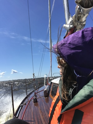 A person wearing a bright orange and green jacket with a purple hood is on a wooden sailboat. The boat is moving through choppy waters under a clear blue sky. There is a sense of motion as the boat cuts through the waves, and the person's hair blows in the wind.