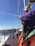 A person wearing a bright orange and green jacket with a purple hood is on a wooden sailboat. The boat is moving through choppy waters under a clear blue sky. There is a sense of motion as the boat cuts through the waves, and the person's hair blows in the wind.