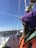 A person wearing a bright orange and green jacket with a purple hood is on a wooden sailboat. The boat is moving through choppy waters under a clear blue sky. There is a sense of motion as the boat cuts through the waves, and the person's hair blows in the wind.