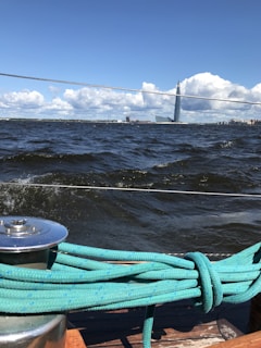 A view from a boat sailing on a body of water with choppy waves. In the foreground, a teal rope is coiled on a metal winch attached to the boat's deck. The background features a distant modern skyscraper tower and several buildings along the coastline. Fluffy white clouds dot the bright blue sky.