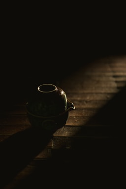 A ceramic teapot is placed on a wooden surface with a dramatic beam of light highlighting its contours. The surrounding area remains dark, creating a strong contrast.