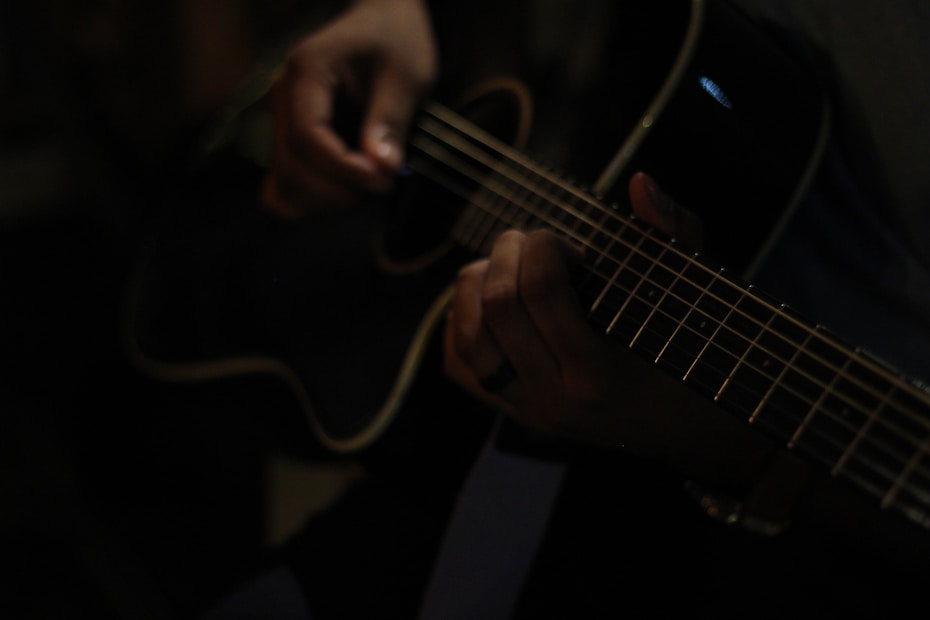 A person is playing an acoustic guitar, focusing on their fingers positioned on the strings. The image is dimly lit, creating a moody atmosphere.