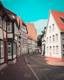 A charming cobblestone street lined with traditional, half-timbered houses featuring white walls and dark wooden beams. The architecture is quaint and historic, with some buildings showcasing red-tiled roofs. A few people can be seen walking down the curved street in the background, under a bright blue sky.