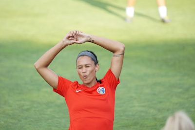 A person in a red sports jersey is standing on a grassy field with their arms raised above their head. The jersey has a logo on the chest. The person appears to be stretching, possibly preparing for athletic activity.