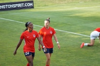 Two soccer players wearing red jerseys and navy shorts are walking on a grass field. One player has the number 20 and the other has number 6 on their shorts. A third player is on the ground in the background. The field is bright and well-maintained.
