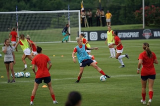 A group of female soccer players engaged in a practice session on a grass field. They wear red and blue uniforms, with some in training bibs. A visible goalpost, cones, and soccer balls are scattered around. In the background, a few spectators watch from sidelines near a banner with logos.