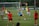 A group of female soccer players engaged in a practice session on a grass field. They wear red and blue uniforms, with some in training bibs. A visible goalpost, cones, and soccer balls are scattered around. In the background, a few spectators watch from sidelines near a banner with logos.