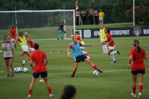 A group of female soccer players engaged in a practice session on a grass field. They wear red and blue uniforms, with some in training bibs. A visible goalpost, cones, and soccer balls are scattered around. In the background, a few spectators watch from sidelines near a banner with logos.