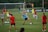 A group of female soccer players engaged in a practice session on a grass field. They wear red and blue uniforms, with some in training bibs. A visible goalpost, cones, and soccer balls are scattered around. In the background, a few spectators watch from sidelines near a banner with logos.